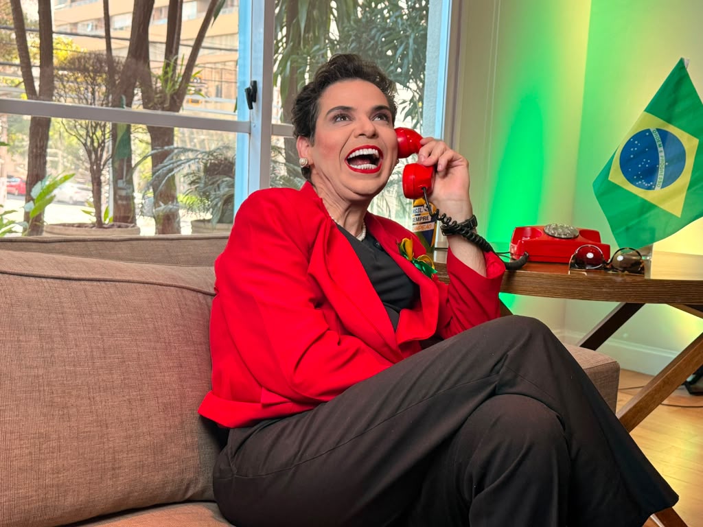 Gustavo Mendes interpretando a personagem Dilma, rindo ao telefone vermelho